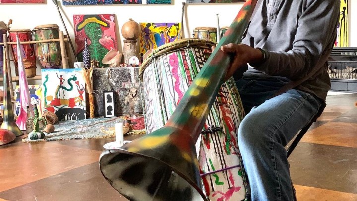 a man plays a spray painted bugle while drumming in a room of colorful paintings