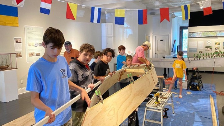 a line of preteen boys participate in building a wooden boat, with the help of a teacher, under a string of maritime flags