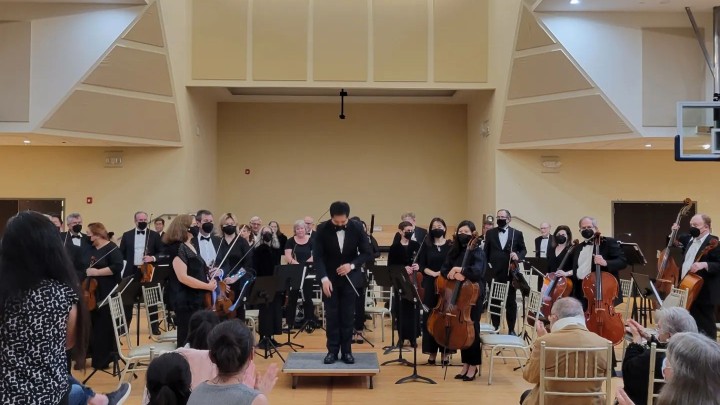 a conductor bows in front of his ensemble while people in yellow chairs clap for him