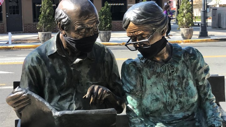 Painted bronze sculpture of two older people reading on a bench titled Steadfast and Loyal on the sidewalk of a town street