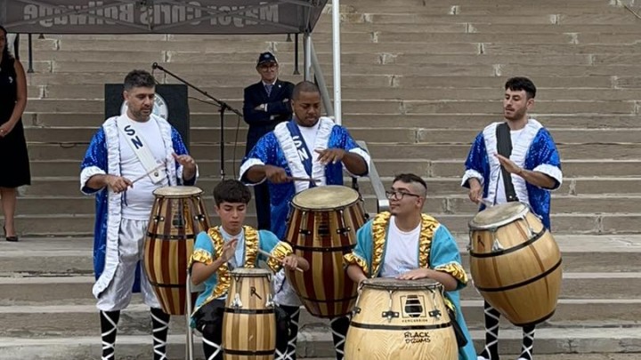 Five men playing drums at the base of stairs leading up to a building.
