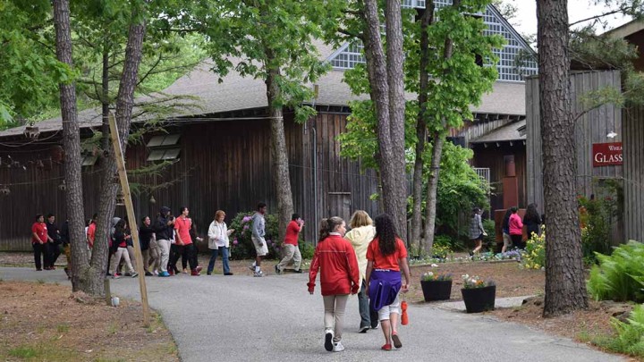 People walking in a wooded area amongst wooden sided buildings at WheatonArts.